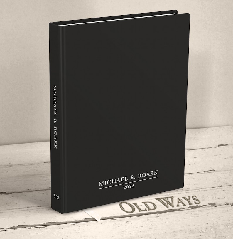 A hardcover casebound book standing upright on a wood surface. The cover of the book is plain black. It is personalized with a name and the year on the cover and spine with white text in a classic serif font.