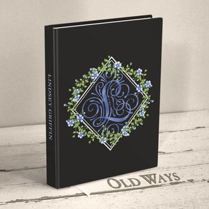 May include: A black hardcover book with the text LINDSEY GRIFFIN on the spine. The front cover features a white diamond frame surrounding a blue calligraphic letter L, decorated with green vines and small blue flowers. The book stands upright.