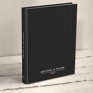 A hardcover casebound book standing upright at an angle on a neutral light wood surface. The cover of the book is minimalist in plain matte black. It is personalized with a name and the year at the bottom of the front cover and spine with white text in a classic serif font.