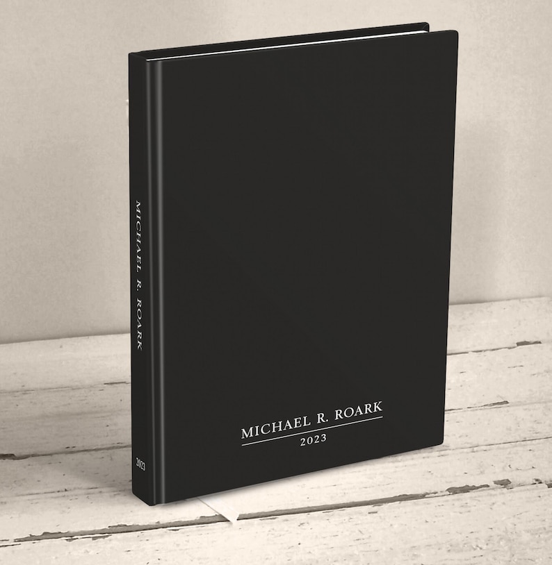A hardcover casebound book standing upright at an angle on a neutral light wood surface. The cover of the book is minimalist in plain matte black. It is personalized with a name and the year at the bottom of the front cover and spine with white text in a classic serif font.