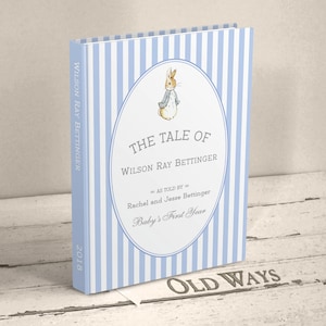 May include: A blue and white striped book cover with a Peter Rabbit illustration. The title is "The Tale of Wilson Ray Bettinger" with the subtitle "As told by Rachel and Jesse Bettinger, Baby's First Year". The year 2018 is printed on the spine.