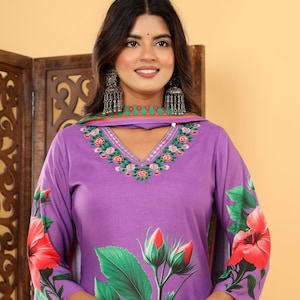 May include: A lilac floral print salwar kameez. The outfit has a V-neck and sleeves decorated with red hibiscus flowers and green leaves. It includes a matching dupatta, a traditional Indian garment.