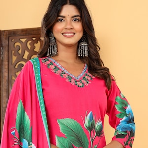 Puede incluir: Un vibrante conjunto kurta con estampado floral rosa. La parte superior presenta un escote en V con detalles bordados y mangas tres cuartos. El dupatta y el kurta a juego están adornados con grandes diseños florales azules y verdes. El conjunto se complementa con pendientes plateados y una pulsera.