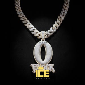 Puede incluir: Un collar con colgante incrustado de diamantes con una gran letra "O" y la palabra "ROCK" debajo, engarzado en una gruesa cadena helada. El colgante y la cadena son de oro y plata, sobre un fondo negro.