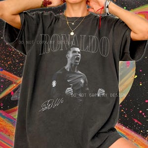 May include: Dark gray oversized t-shirt featuring a black and white image of a soccer player with the name "RONALDO" in large white letters. The shirt also includes a signature and the text "DO NOT COPY MY DESIGN".