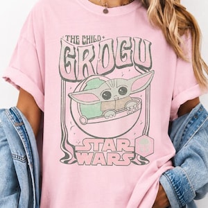 May include: A light pink short-sleeve t-shirt featuring a graphic print of the character Grogu from Star Wars. The design includes the text THE CHILD GROGU and STAR WARS in a stylized font, with a blue denim jacket layered over the shoulders.