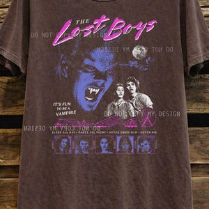 May include: Espresso-colored t-shirt featuring a graphic from "The Lost Boys" movie. The design includes a vampire face, the movie title in pink, and the phrase "It's Fun to Be a Vampire." The shirt also has the text "Sleep All Day - Party All Night - Never Grow Old - Never Die."