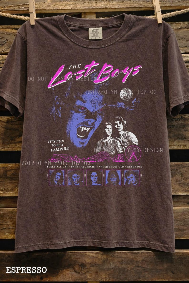 May include: Espresso-colored t-shirt featuring a graphic from "The Lost Boys" movie. The design includes a vampire face, the movie title in pink, and the phrase "It's Fun to Be a Vampire." The shirt also has the text "Sleep All Day - Party All Night - Never Grow Old - Never Die."