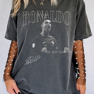 May include: Dark gray t-shirt featuring the name "RONALDO" in white above a black and white image of a soccer player. The shirt includes the player's signature and a small logo. The shirt has a vintage, worn look.