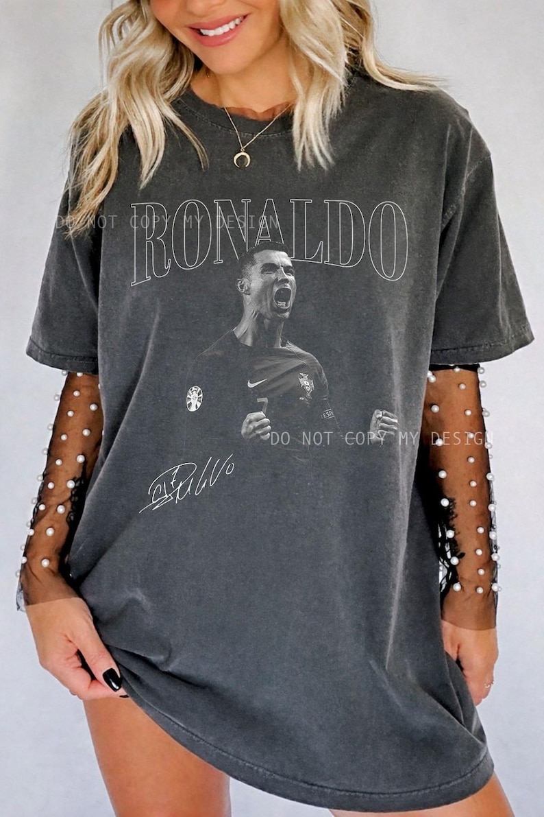 May include: Dark gray t-shirt featuring the name "RONALDO" in white above a black and white image of a soccer player. The shirt includes the player's signature and a small logo. The shirt has a vintage, worn look.