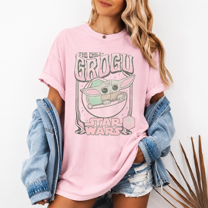 May include: A light pink short-sleeve t-shirt featuring a graphic print of the character Grogu from Star Wars. The design includes the text THE CHILD GROGU and STAR WARS in a stylized font, with a blue denim jacket layered over the shoulders.