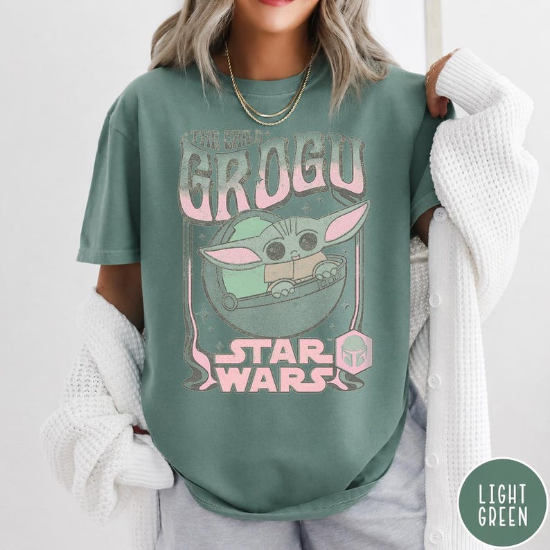 May include: A sage green short-sleeve t-shirt featuring a graphic print of Grogu in a floating pod. The design includes the text GROGU and STAR WARS in a light pink, distressed font. The shirt is worn with a white, textured, long-sleeve cardigan.