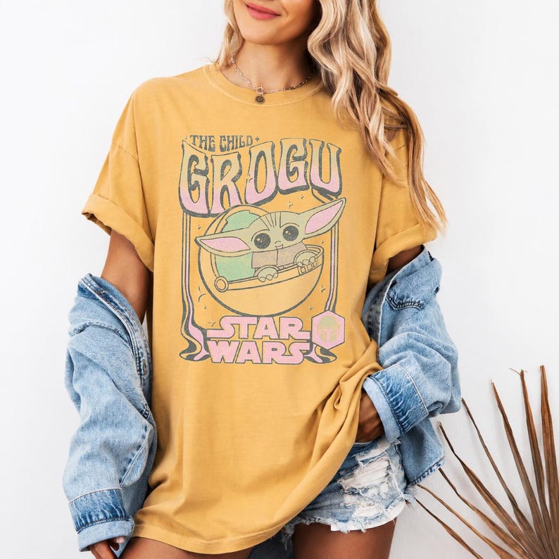 May include: A mustard yellow short-sleeve t-shirt featuring a graphic print of Grogu from Star Wars. The design includes the text THE CHILD GROGU and STAR WARS in pink and white lettering, with a circular illustration of the character in a pod.