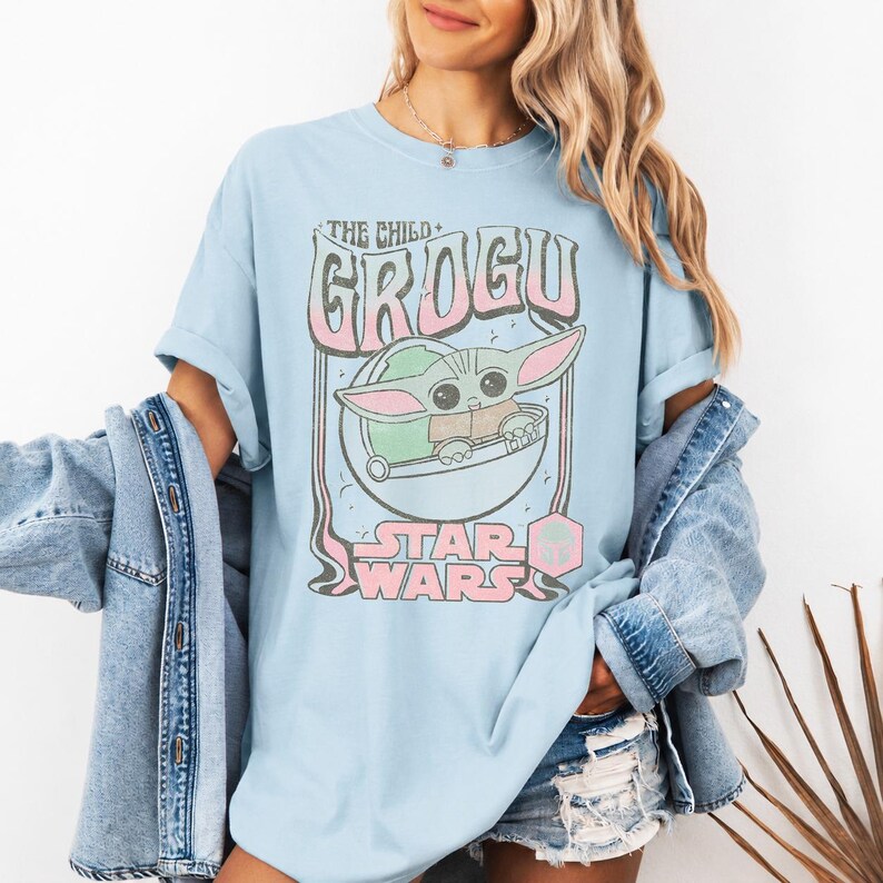 May include: A light blue t-shirt featuring a graphic print of the character Grogu from Star Wars. The design includes the text THE CHILD GROGU and STAR WARS in a retro font style, with pink and green accents, layered over a denim jacket.