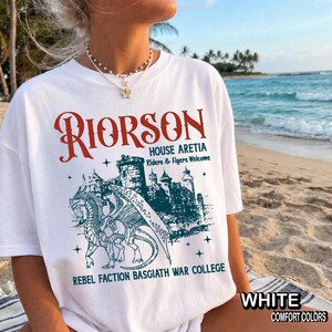 Puede incluir: Camiseta blanca con un dise&ntilde;o de fantas&iacute;a. El dise&ntilde;o presenta la palabra "RIORSON" en rojo, una ilustraci&oacute;n de un castillo y el texto "HOUSE ARETIA, Riders & Flyers Welcome, REBEL FACTION BASGIATH WAR COLLEGE". La camiseta est&aacute; etiquetada como "WHITE COMFORT COLORS".