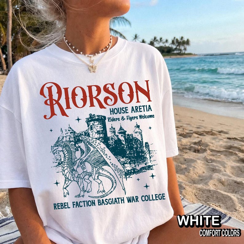Puede incluir: Camiseta blanca con un dise&ntilde;o de fantas&iacute;a. El dise&ntilde;o presenta la palabra "RIORSON" en rojo, una ilustraci&oacute;n de un castillo y el texto "HOUSE ARETIA, Riders & Flyers Welcome, REBEL FACTION BASGIATH WAR COLLEGE". La camiseta est&aacute; etiquetada como "WHITE COMFORT COLORS".