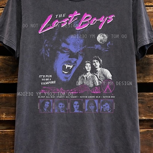 May include: Dark gray t-shirt featuring a graphic from "The Lost Boys" movie. The design includes a vampire, two people, and the text "The Lost Boys", "It's fun to be a vampire", and "Sleep all day, party all night, never grow old, never die."