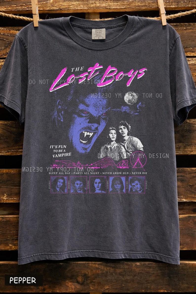 May include: Dark gray t-shirt featuring a graphic from "The Lost Boys" movie. The design includes a vampire, two people, and the text "The Lost Boys", "It's fun to be a vampire", and "Sleep all day, party all night, never grow old, never die."