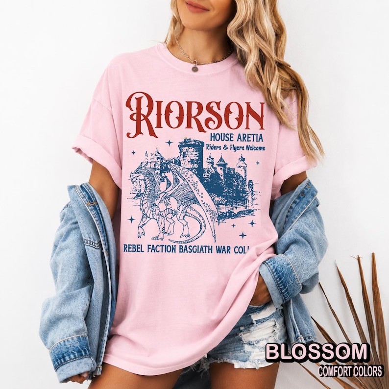 Puede incluir: Camiseta rosa claro con un dise&ntilde;o gr&aacute;fico que presenta un drag&oacute;n, un castillo y texto. El texto incluye "RIORSON", "HOUSE ARETIA" y "REBEL FACTION BASGIATH WAR COL". La camiseta tambi&eacute;n tiene la palabra "BLOSSOM" y "COMFORT COLORS".
