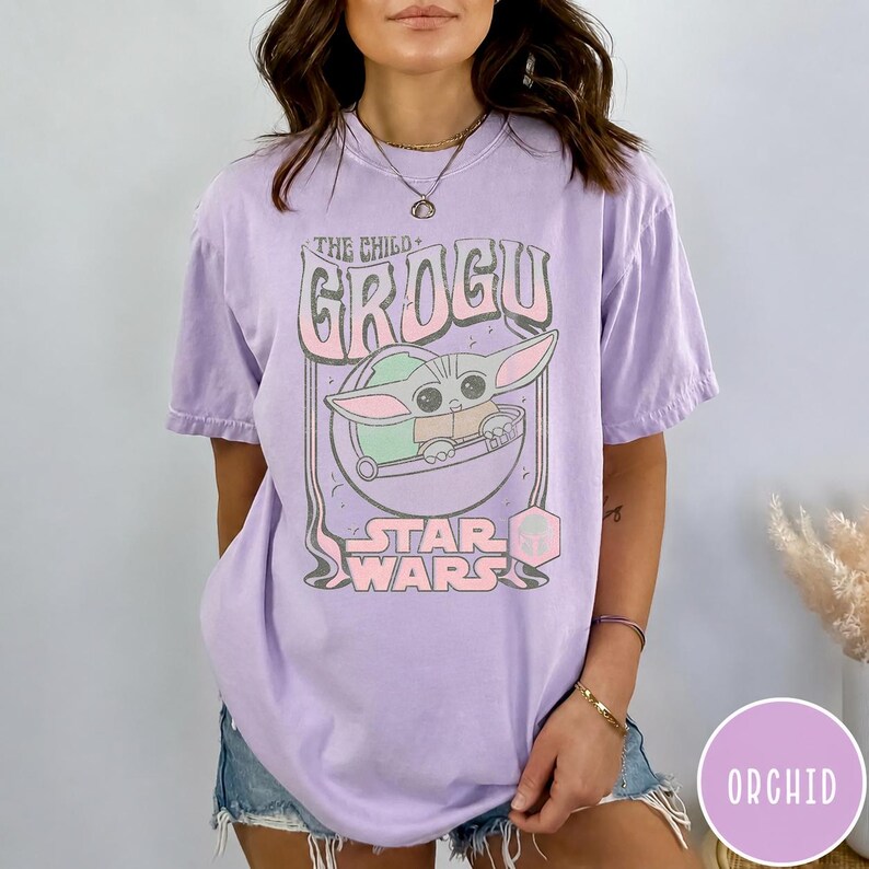 May include: A light purple short-sleeve t-shirt featuring a graphic print of the character Grogu from Star Wars. The design includes the text THE CHILD GROGU and STAR WARS in a stylized font, with a circular frame around the character illustration.