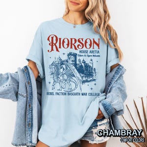 Puede incluir: Camiseta azul claro con un dise&ntilde;o de drag&oacute;n y castillo, y texto que dice "RIORSON HOUSE ARETIA Riders & Figers Welcome REBEL FACTION BASGIATH WAR COLLEGE". La camiseta tambi&eacute;n tiene la palabra "CHAMBRAY" y "COMFORT COLORS".