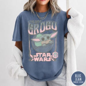 May include: A blue short-sleeve t-shirt featuring a graphic print of Grogu in a floating pod. The text reads THE CHILD GROGU and STAR WARS in a distressed pink and white font. The design includes small star accents and a helmet icon.