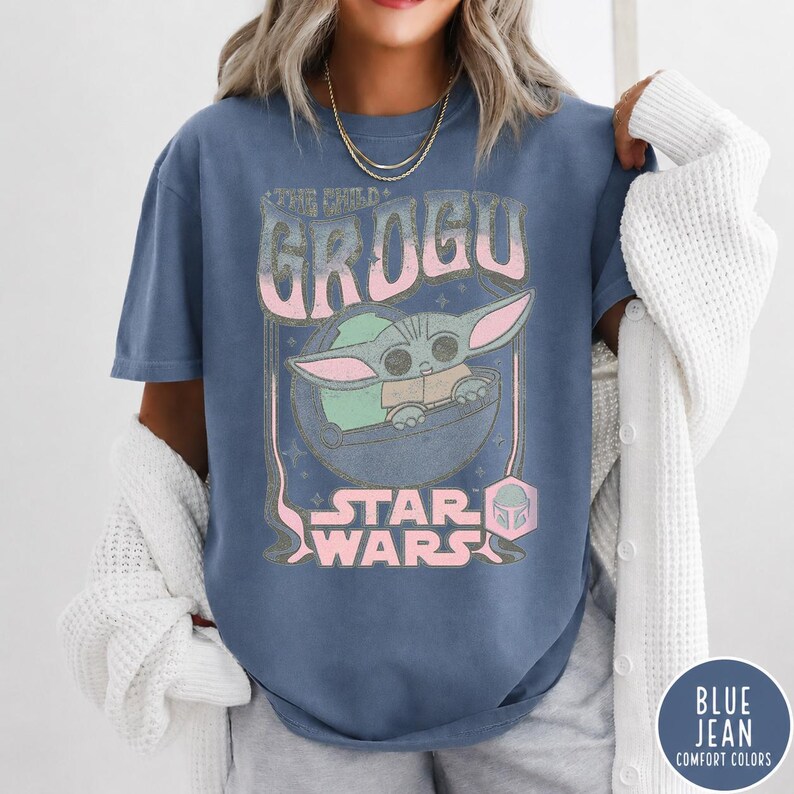 May include: A blue short-sleeve t-shirt featuring a graphic print of Grogu in a floating pod. The text reads THE CHILD GROGU and STAR WARS in a distressed pink and white font. The design includes small star accents and a helmet icon.