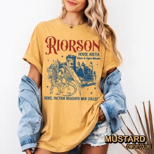 Puede incluir: Camiseta de color mostaza con un gr&aacute;fico que presenta el texto "RIORSON" y una ilustraci&oacute;n detallada de un drag&oacute;n y un castillo. El texto adicional incluye "HOUSE ARETIA" y "REBEL FACTION BASGIATH WAR COLLEG". La camiseta est&aacute; etiquetada como "MUSTARD" y "COMFORT COLORS".