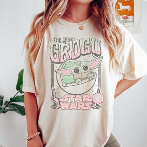 May include: A cream-colored t-shirt featuring a graphic print of Grogu from Star Wars. The design includes the text THE CHILD, GROGU, and STAR WARS in a retro font, with a pink and green color palette. A small pink hexagonal icon is also visible.