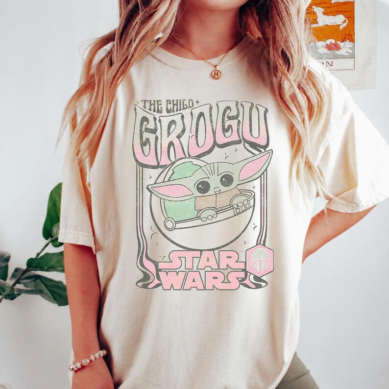 May include: A cream-colored t-shirt featuring a graphic print of Grogu from Star Wars. The design includes the text THE CHILD, GROGU, and STAR WARS in a retro font, with a pink and green color palette. A small pink hexagonal icon is also visible.