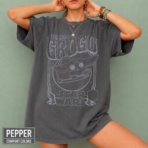May include: A charcoal gray short-sleeve t-shirt featuring a light gray graphic print of Grogu from Star Wars inside a circular frame. The text reads THE CHILD GROGU and STAR WARS. The shirt has a faded, washed-out appearance and a crew neckline.