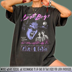 May include: Dark gray t-shirt featuring a graphic from "The Lost Boys" movie. The design includes a vampire illustration, the movie title, and the phrase "It's Fun To Be A Vampire." Additional text includes "Sleep All Day, Party All Night, Never Grow Old, Never Die."