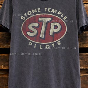 K&ouml;nnte beinhalten: Dunkelgraues T-Shirt mit dem Text "STONE TEMPLE PILOTS" und "STP" in einem rot-wei&szlig;en Oval. Das Shirt hat einen Vintage-Look und h&auml;ngt vor einem Holzuntergrund.