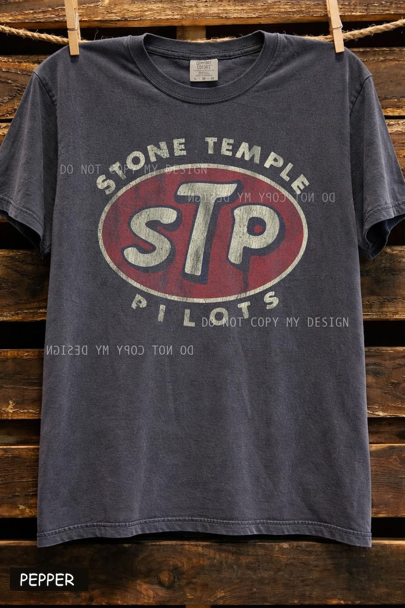 K&ouml;nnte beinhalten: Dunkelgraues T-Shirt mit dem Text "STONE TEMPLE PILOTS" und "STP" in einem rot-wei&szlig;en Oval. Das Shirt hat einen Vintage-Look und h&auml;ngt vor einem Holzuntergrund.