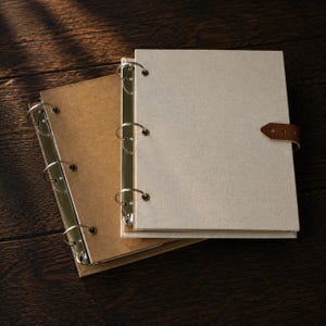 Può includere: Due raccoglitori a tre anelli con meccanismi in metallo. Un raccoglitore presenta una copertina in tessuto beige chiaro, mentre l'altro ha una copertina in carta marrone. Il raccoglitore beige include una chiusura a cinturino in similpelle.