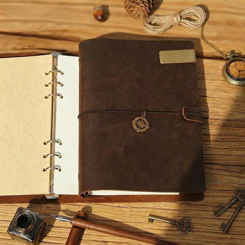 May include: A brown leather journal with a brass clasp and a compass charm. The journal is open, revealing blank pages. A vintage pocket watch, keys, and inkwell are also present, suggesting a theme of travel and writing.