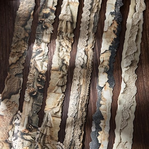 Puede incluir: Primer plano de varias tiras de papel decorativo con una estética vintage. Las tiras presentan bordes rasgados, detalles de encaje y texto impreso, en tonos marrón, beige y negro. Las tiras de papel están dispuestas verticalmente sobre un fondo de madera oscura.