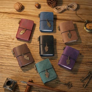 Op de afbeelding: Een verzameling kleine, leren notitieboekjes in diverse kleuren, waaronder bordeauxrood, blauw, bruin, zwart, groen en paars. Elk notitieboekje heeft een decoratieve bladbedel en een zwarte elastische sluiting. De notitieboekjes liggen op een houten oppervlak.