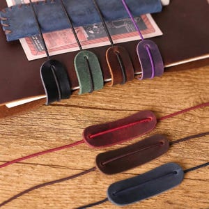 May include: A collection of leather journal bookmarks in various colors, including black, green, brown, and purple. Each bookmark has a loop and a colored cord. The bookmarks are arranged on a wooden surface.