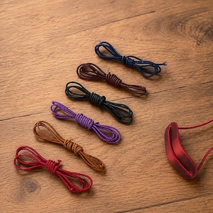 May include: A collection of six thin leather cords in various colours including navy blue, dark brown, black, purple, tan, and red. Each cord is neatly coiled into a small loop. A single red leather component with a curved slot is also visible.