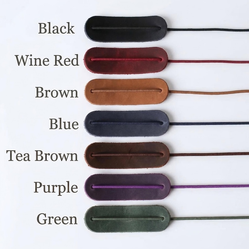 May include: A selection of leather cord keepers in various colors, including black, wine red, brown, blue, tea brown, purple, and green. Each keeper is an oval shape with a cord attached, displayed against a white background.