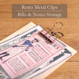 Puede incluir: Clip retro de metal bronce que sujeta papel vintage con una ilustración histórica y texto. El clip sujeta el papel de forma segura. El papel presenta la palabra "Enero" y texto sobre la "Revolución".
