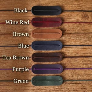 May include: A product display featuring seven leather cord keepers in various colors: black, wine red, brown, blue, tea brown, purple, and green. Each keeper has a slot for a cord and is displayed on a wooden surface. The colors are labeled.