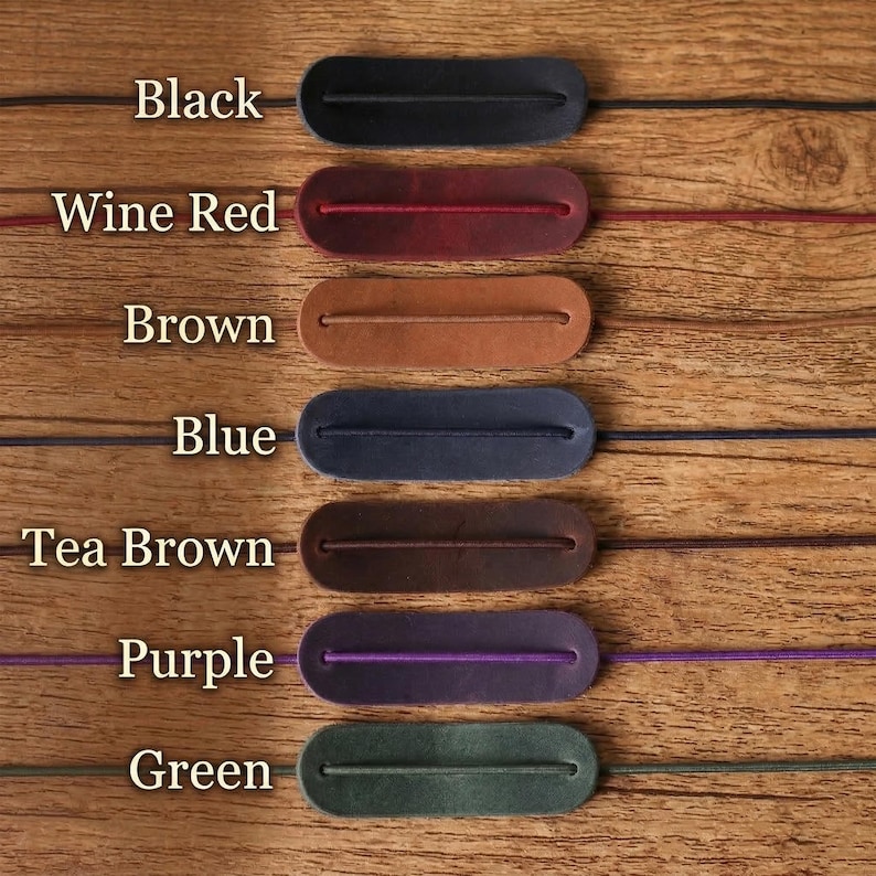 May include: A product display featuring seven leather cord keepers in various colors: black, wine red, brown, blue, tea brown, purple, and green. Each keeper has a slot for a cord and is displayed on a wooden surface. The colors are labeled.