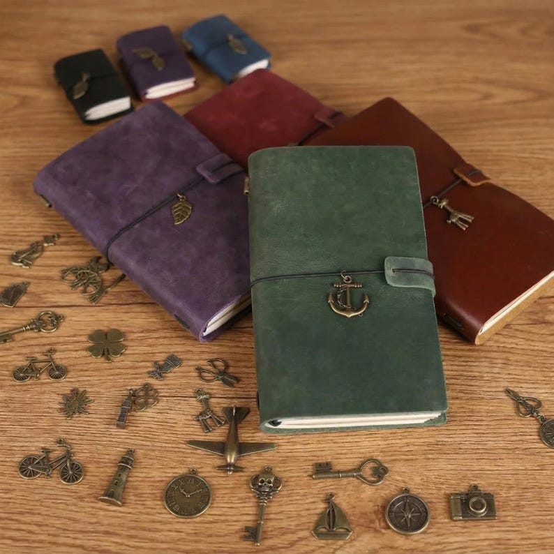 May include: A collection of leather-bound journals in various colors, including purple, green, and brown, with decorative metal charms scattered around. The journals have elastic closures and metal accents. The charms include keys, bicycles, and other vintage-style designs.