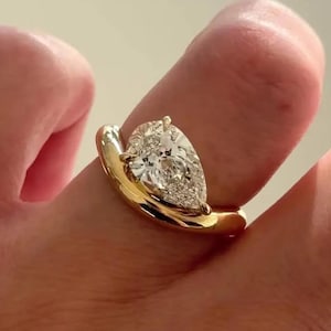 Peut inclure: Une bague en or ornée d'un diamant en forme de poire. Le corps de la bague épouse le doigt avec une forme incurvée. Le diamant scintille, reflétant la lumière. Il s'agit d'un bijou.