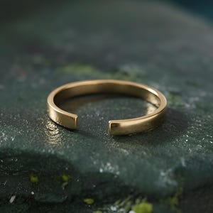 Op de afbeelding: Een goudkleurige, open ring. De ring ligt op een donkergroen, getextureerd oppervlak. Het ontwerp van de ring heeft een eenvoudige, minimalistische stijl, met een kleine opening aan de voorkant.