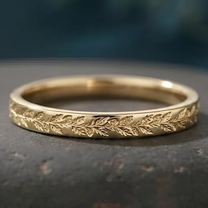 Może przedstawiać: Złoty pierścionek z ciągłym grawerowanym wzorem delikatnych liści i pnączy wokół całej zewnętrznej obrączki. Metal ma polerowane wykończenie, które odbija światło, podkreślając misterną roślinną fakturę wyrytą na powierzchni przedmiotu.