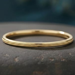 May include: A gold ring with a beaded design around the band. The ring is set against a dark, textured background. The ring is a simple band style, suitable for everyday wear or as a wedding band.