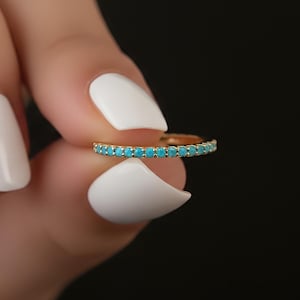 May include: A gold-colored ring with a row of small, bright turquoise stones. The ring is held between fingers with white nail polish against a black background. The ring is a piece of jewelry.